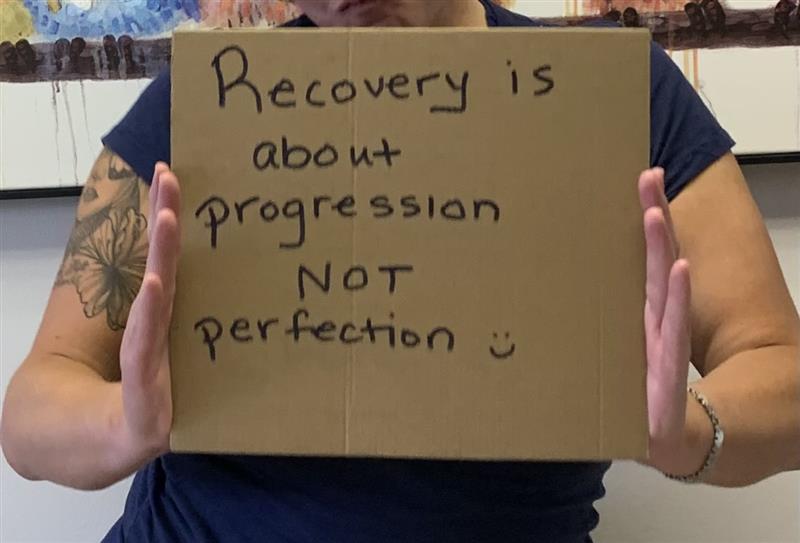 A person holding a cardboard sign that says Recovery is about progression not perfection